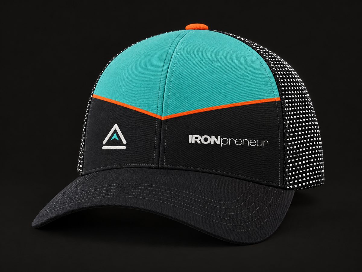 Concept render: the IronPreneur trucker cap in three-quarter view, teal crown, ember V piping, charcoal front with the chevron icon and IRONpreneur wordmark, black dotted mesh.